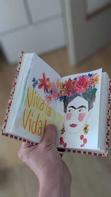 Das Bild zeigt eine farbenfrohe Illustration der Künstlerin Frida Kahlo. Neben der Fogur stehet ein Schriftzug: Viva la Vida!