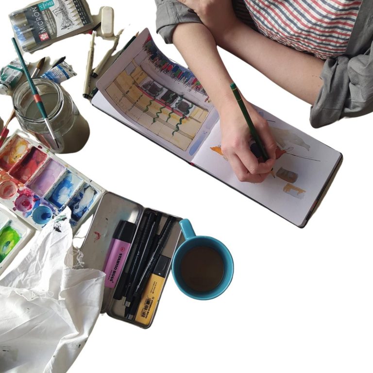 An einem Tisch mit vielen verschiedenen Künstler-Utensilien, von oben aufgenommen sieht man die rechte Hand der Künstlerin. Sie hält einen Pinsel und malt in ein Skizzenbuch.